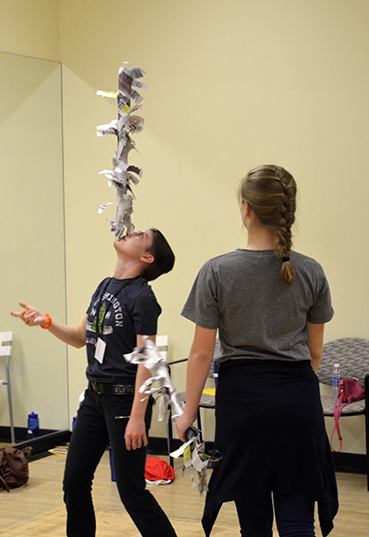 A boy balances a newspaper sculpture on his chin while a girl watches.