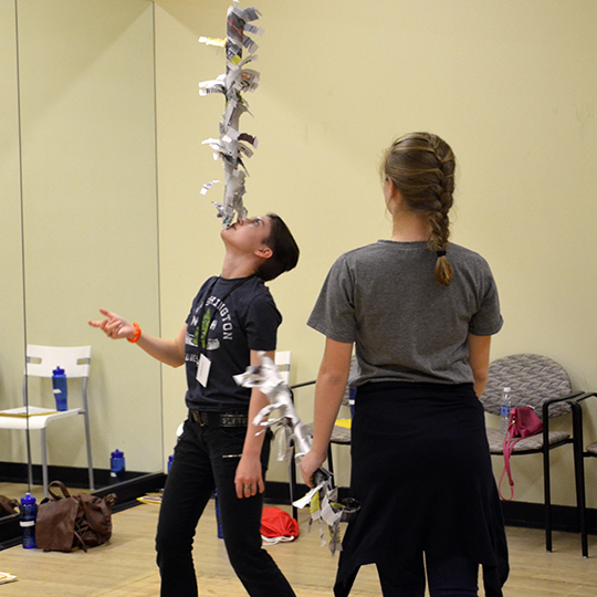 A boy balances a newspaper sculpture on his chin while a girl watches.
